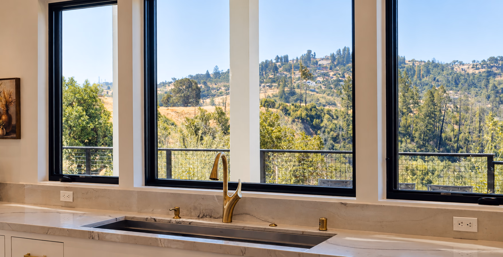Brass faucet and undermount sink under windows onto wooded hills